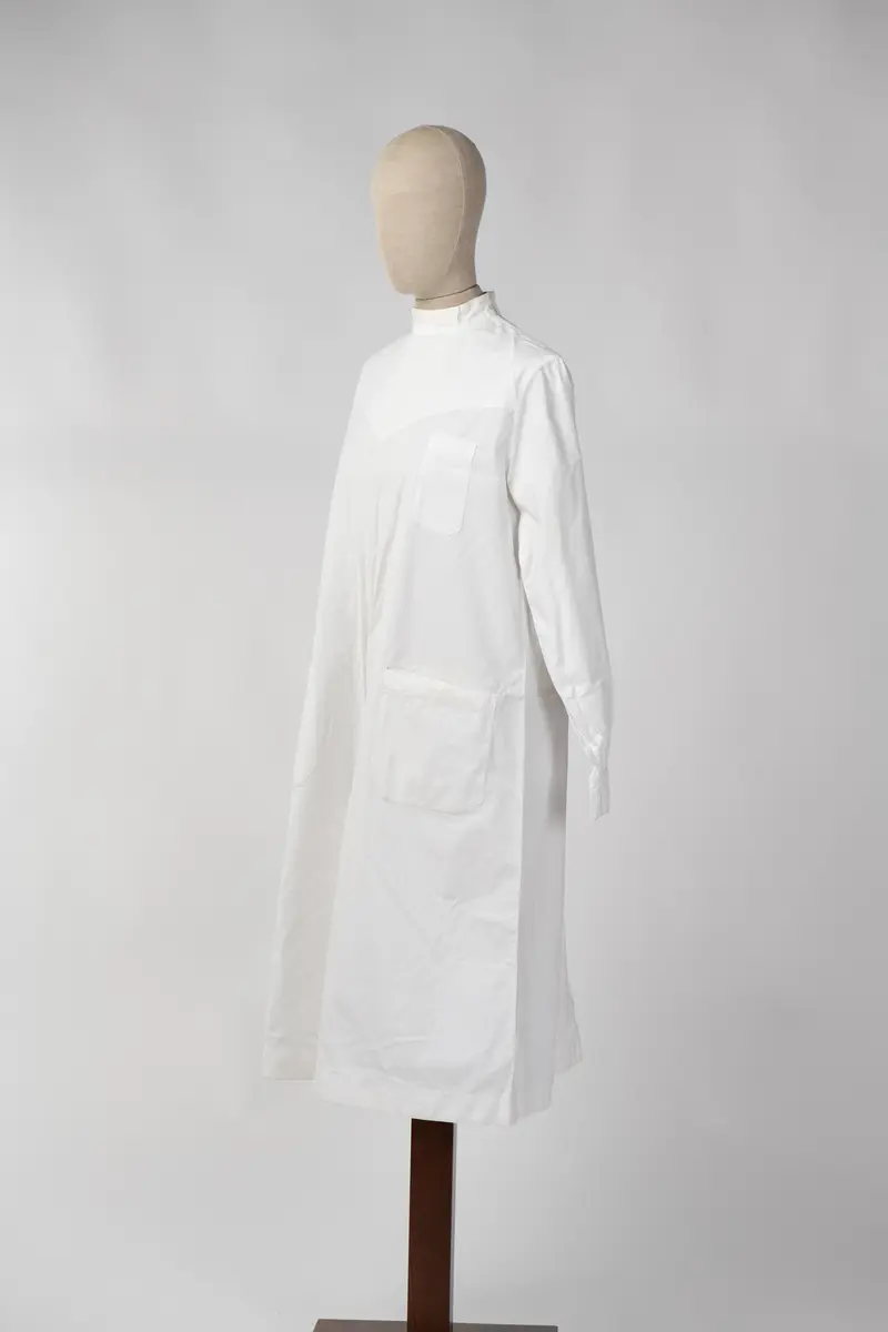 Lang sykepleieruniform i hvit bomull. Uniformen har høy hals med 4 knapper som går halvveis ned på ryggen, og manskjett på ermene med 1 knapp. På forsiden er det 1 liten lomme øverst på venstre side og 2 større lommer på midten av uniformen.