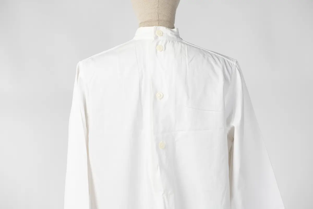 Lang sykepleieruniform i hvit bomull. Uniformen har høy hals med 4 knapper som går halvveis ned på ryggen, og manskjett på ermene med 1 knapp. På forsiden er det 1 liten lomme øverst på venstre side og 2 større lommer på midten av uniformen.