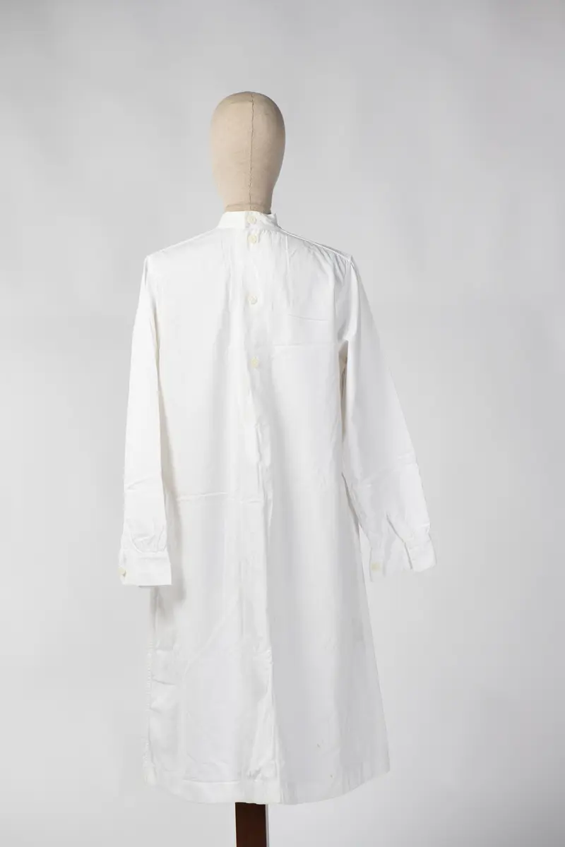 Lang sykepleieruniform i hvit bomull. Uniformen har høy hals med 4 knapper som går halvveis ned på ryggen, og manskjett på ermene med 1 knapp. På forsiden er det 1 liten lomme øverst på venstre side og 2 større lommer på midten av uniformen.