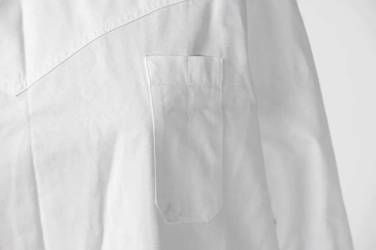 Lang sykepleieruniform i hvit bomull. Uniformen har høy hals med 4 knapper som går halvveis ned på ryggen, og manskjett på ermene med 1 knapp. På forsiden er det 1 liten lomme øverst på venstre side og 2 større lommer på midten av uniformen.