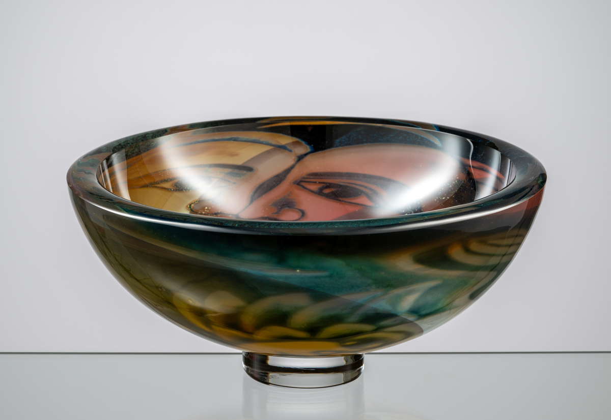 Formgivare: Eva Englund. Graalskål, tjockväggig rund form med klar fotklack. Figurmotiv, tre ansikten varav en ängel i beige, grön och blekrosa nyans mot mörk fond.