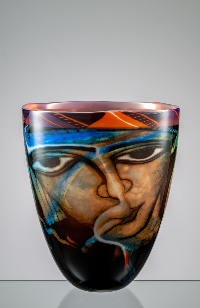 Formgivare: Eva Englund. Graalvas, ovoid form med motiv av två ansikten i blekröd, beige och blågrön nyans mot mörk fond. Blekröd mönstrad bård runt mynningen.