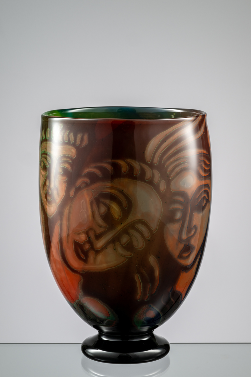 Formgivare: Eva Englund. Graalvas, ovoid form med svart låg fot. Figurmotiv med flertal ansikten i beige, grön och blekrosa nyans mot mörk fond.