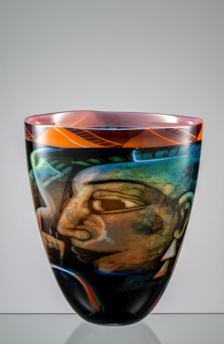 Formgivare: Eva Englund. Graalvas, ovoid form med motiv av två ansikten i blekröd, beige och blågrön nyans mot mörk fond. Blekröd mönstrad bård runt mynningen.