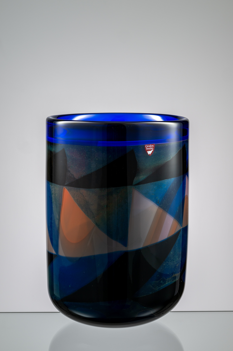 Formgivare: Eva Englund. Graalvas, cylindrisk form med rundad bas. Grafisk dekor i dova nyanser av blått, blekrosa, svagt rött och svart. Blå bård runt mynningen.