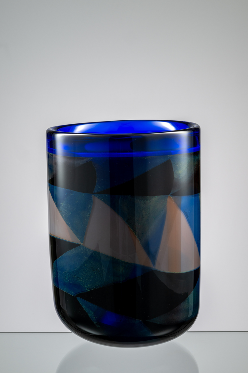 Formgivare: Eva Englund. Graalvas, cylindrisk form med rundad bas. Grafisk dekor i dova nyanser av blått, blekrosa, svagt rött och svart. Blå bård runt mynningen.