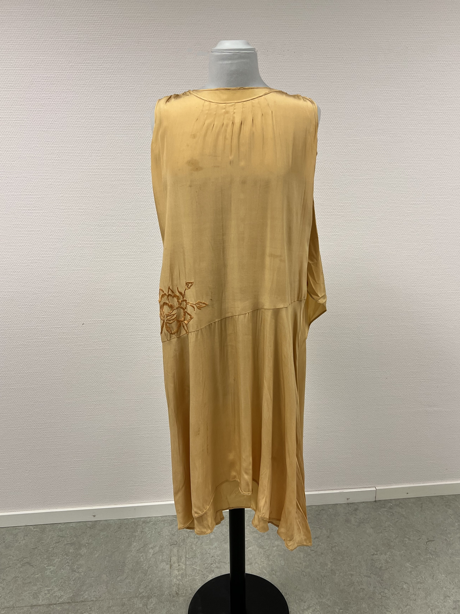 Gul ärmlös klänning med dekoration (schalliknande på ena axeln). Broderad blomma i gult på ena höften. Rundad hals. Sydd med låg midj.