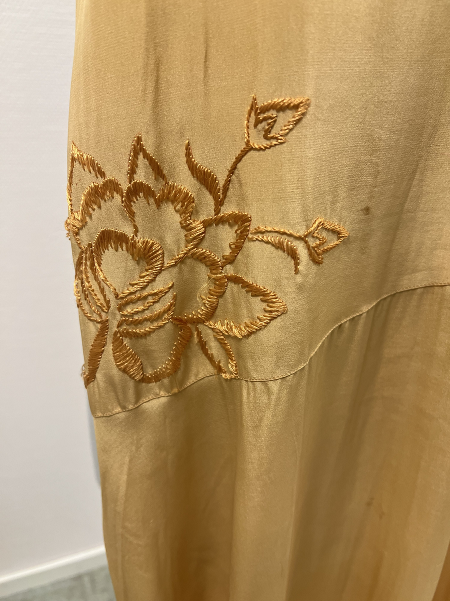 Gul ärmlös klänning med dekoration (schalliknande på ena axeln). Broderad blomma i gult på ena höften. Rundad hals. Sydd med låg midj.