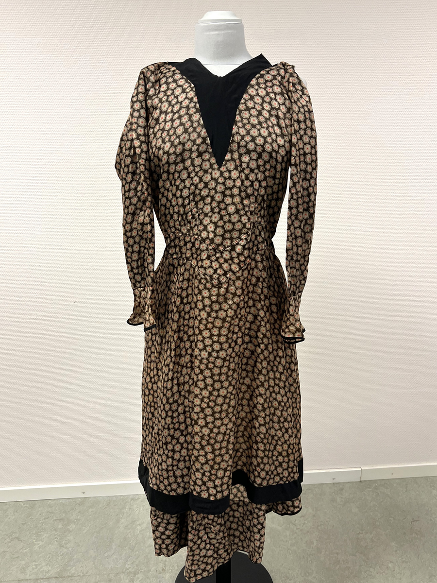 Mönstrad brun, beige, röd och svart klänning. V-ringad hals, svart infälld v-ringning vid hals. Kantad i svart vid ärmmudd och nedtill vid kjolens fåll, I nacken fastsytt band som hänger ned i ryggen, längst ner på banden en förlängning med svarta pärlor och en stor och några mindre guldmålade pärlor i trä. Längre kjol i bakre delen av kjolen.