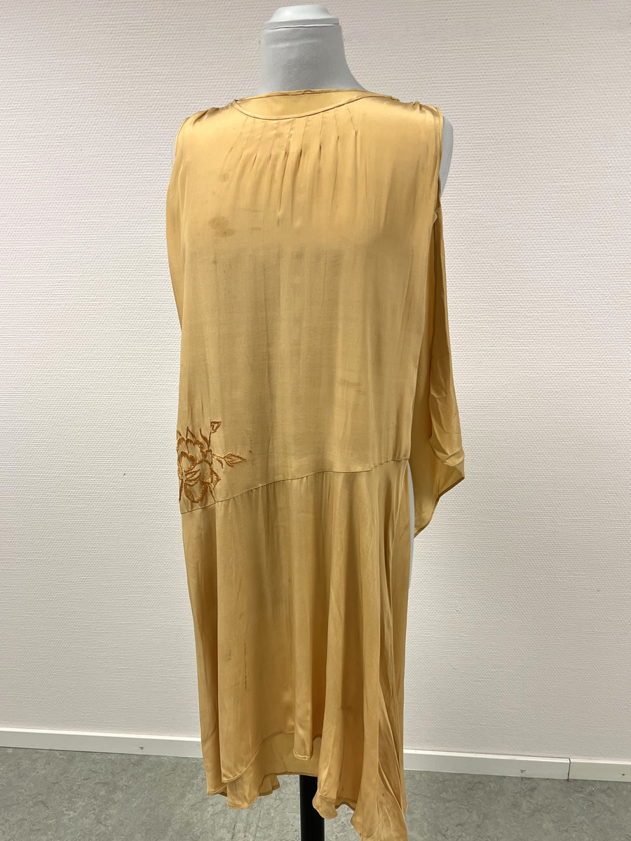 Gul ärmlös klänning med dekoration (schalliknande på ena axeln). Broderad blomma i gult på ena höften. Rundad hals. Sydd med låg midj.