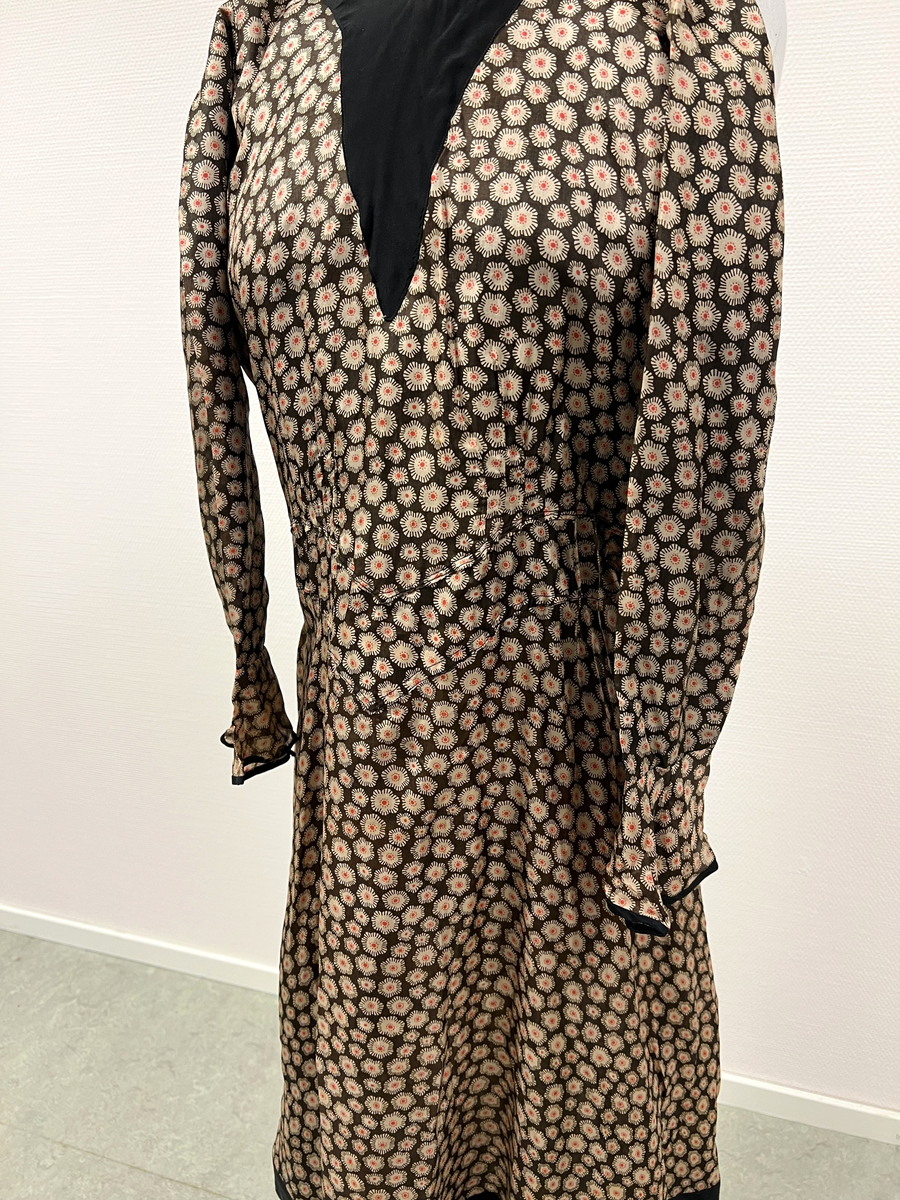 Mönstrad brun, beige, röd och svart klänning. V-ringad hals, svart infälld v-ringning vid hals. Kantad i svart vid ärmmudd och nedtill vid kjolens fåll, I nacken fastsytt band som hänger ned i ryggen, längst ner på banden en förlängning med svarta pärlor och en stor och några mindre guldmålade pärlor i trä. Längre kjol i bakre delen av kjolen.