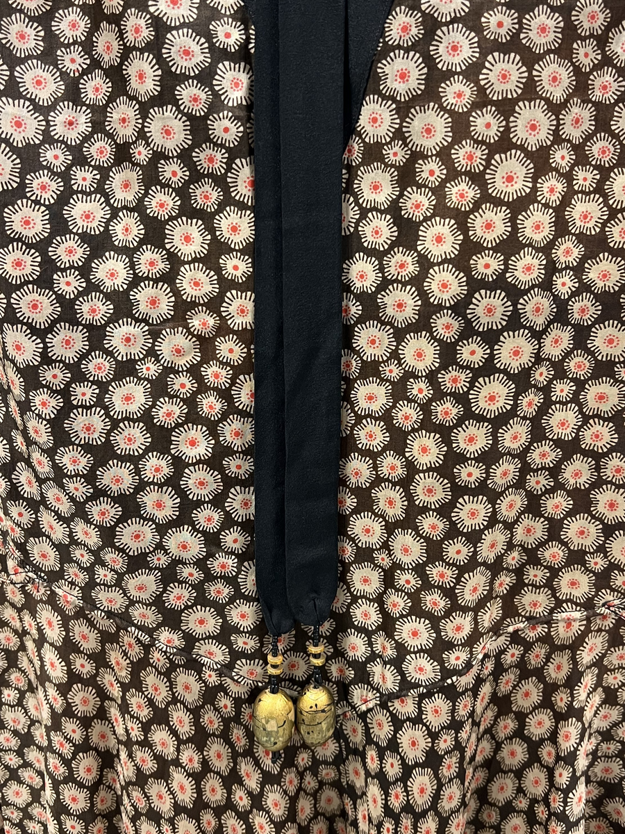 Mönstrad brun, beige, röd och svart klänning. V-ringad hals, svart infälld v-ringning vid hals. Kantad i svart vid ärmmudd och nedtill vid kjolens fåll, I nacken fastsytt band som hänger ned i ryggen, längst ner på banden en förlängning med svarta pärlor och en stor och några mindre guldmålade pärlor i trä. Längre kjol i bakre delen av kjolen.