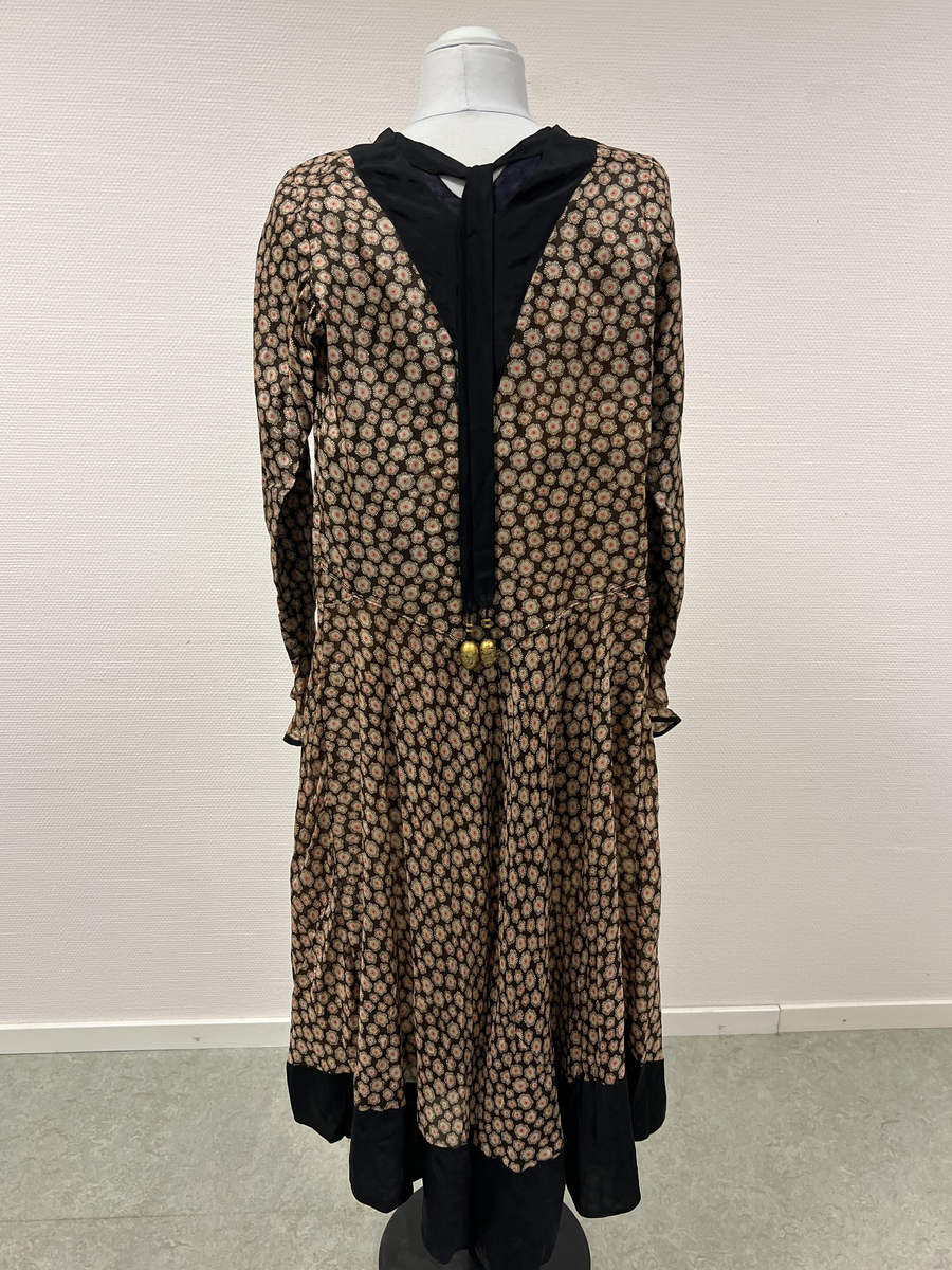 Mönstrad brun, beige, röd och svart klänning. V-ringad hals, svart infälld v-ringning vid hals. Kantad i svart vid ärmmudd och nedtill vid kjolens fåll, I nacken fastsytt band som hänger ned i ryggen, längst ner på banden en förlängning med svarta pärlor och en stor och några mindre guldmålade pärlor i trä. Längre kjol i bakre delen av kjolen.