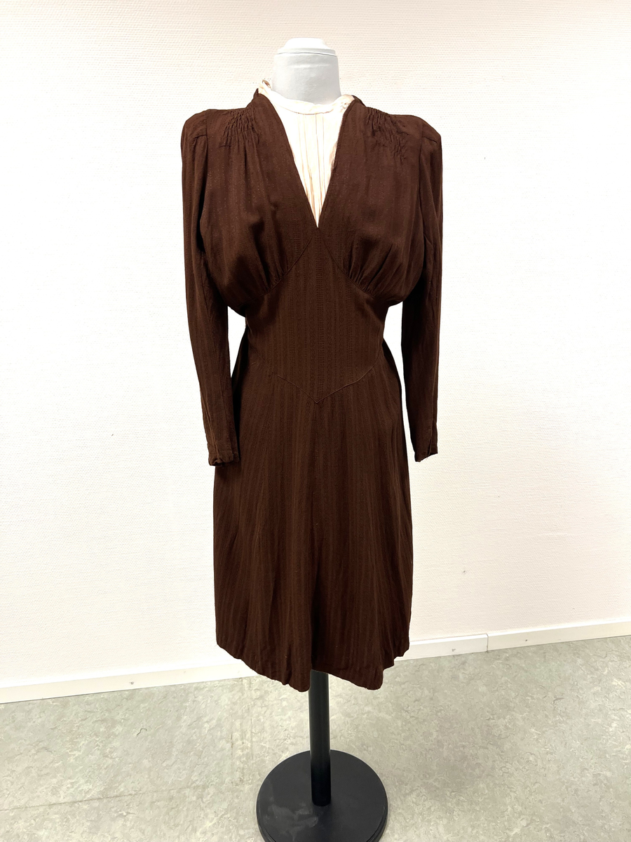 Rödbrun klännning med isättning av ljust tyg vid halsen. Lätt plisserad från axlarna och en liten bit ner på framstycke. Klädda knappar i nacken, men stängs med tryckknappar. Även klädda knappar vid handleden på ärmen (6 st/ärm), men stängs även där med tryckknappar. Kjolen lätt utsvängd. Sleif i ryggen, som på ena sidan är fastsydd och på den andra fastsatt med tryckknappar.