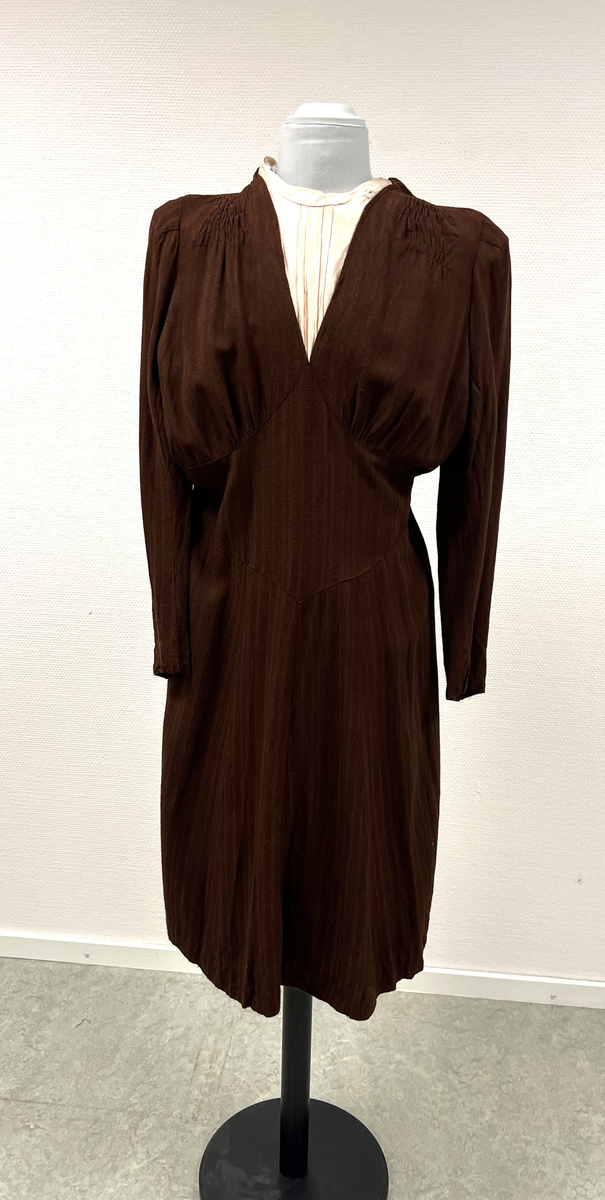 Rödbrun klännning med isättning av ljust tyg vid halsen. Lätt plisserad från axlarna och en liten bit ner på framstycke. Klädda knappar i nacken, men stängs med tryckknappar. Även klädda knappar vid handleden på ärmen (6 st/ärm), men stängs även där med tryckknappar. Kjolen lätt utsvängd. Sleif i ryggen, som på ena sidan är fastsydd och på den andra fastsatt med tryckknappar.