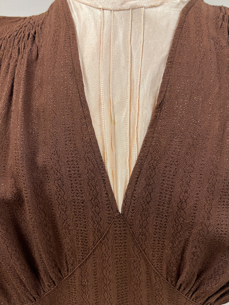 Rödbrun klännning med isättning av ljust tyg vid halsen. Lätt plisserad från axlarna och en liten bit ner på framstycke. Klädda knappar i nacken, men stängs med tryckknappar. Även klädda knappar vid handleden på ärmen (6 st/ärm), men stängs även där med tryckknappar. Kjolen lätt utsvängd. Sleif i ryggen, som på ena sidan är fastsydd och på den andra fastsatt med tryckknappar.