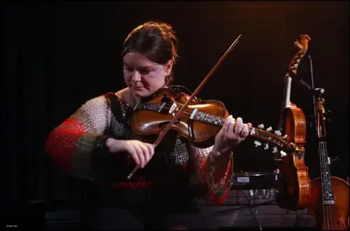 Hannah Moira Midtbø Bruner står på ei scene og spelar hardingfele med fleire hardingfeler i bakgrunnen hengande på eit stativ