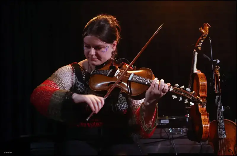 Hannah Moira Midtbø Bruner står på ei scene og spelar hardingfele med fleire hardingfeler i bakgrunnen hengande på eit stativ