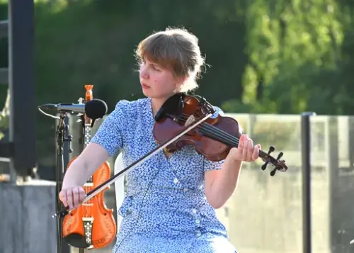 Sunniva L. Røiland i blå og kvit blomstrete kjole sit på ei utescene og spelar på ein fiolin