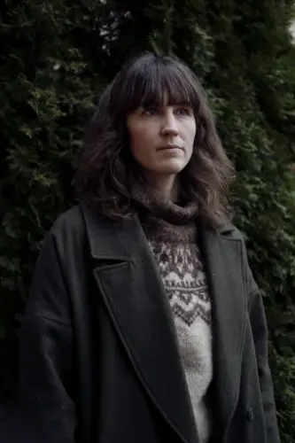 Runa Kvalsund med strikkegenser og mørk frakk står ute framom ein grøn hekk