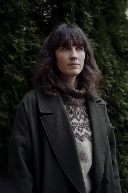 Runa Kvalsund med strikkegenser og mørk frakk står ute framom ein grøn hekk