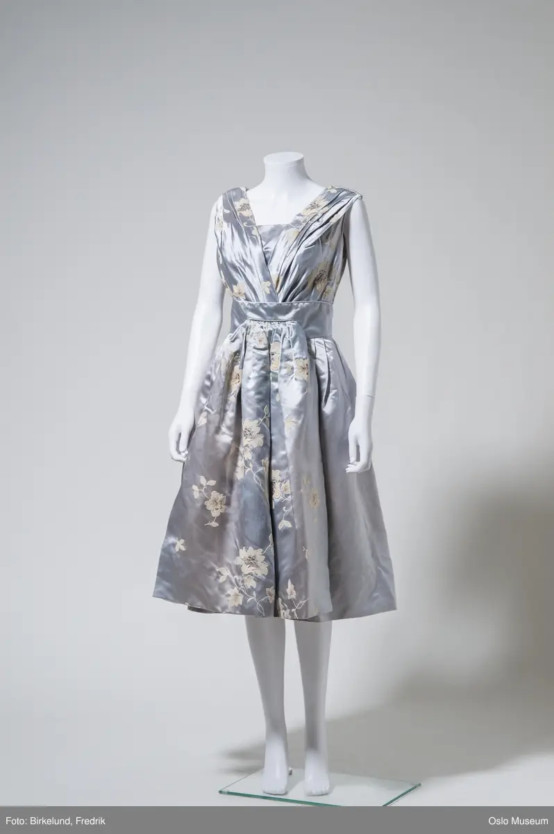 Sølvfarget selskapskjole med utsvingt skjørt og skulderstropper. Lukkes i sdien med glidelås. Dekorert med maskinboderte blomster i hvitt og gull. Forkle festes med hekter.