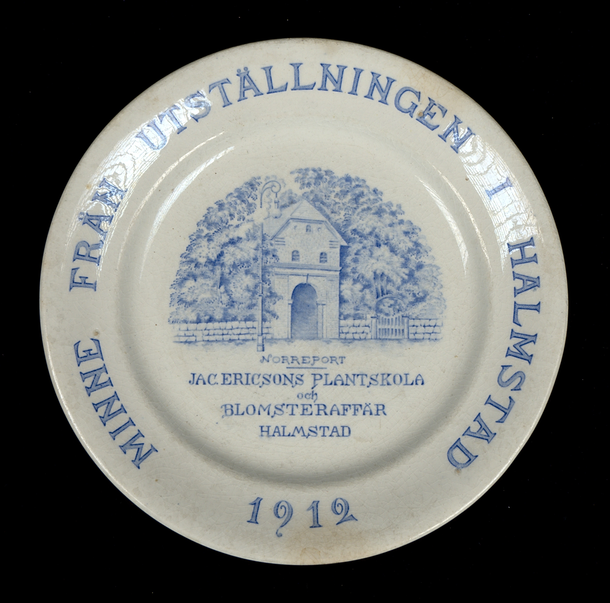 Prydnadsfat av flintgods med tryckt motiv i blått. Längs brämet löper texten: "MINNE FRÅN UTSTÄLLNINGEN I HALMSTAD 1912". I fatets spegel avbildas Norreport i Halmstad, samt texten: "JAC.ERICSONS PLANTSKOLA och BLOMSTERAFFÄR HALMSTAD". Undersidan är stämplad "Rörstrand".

Glasyr krackelerad.