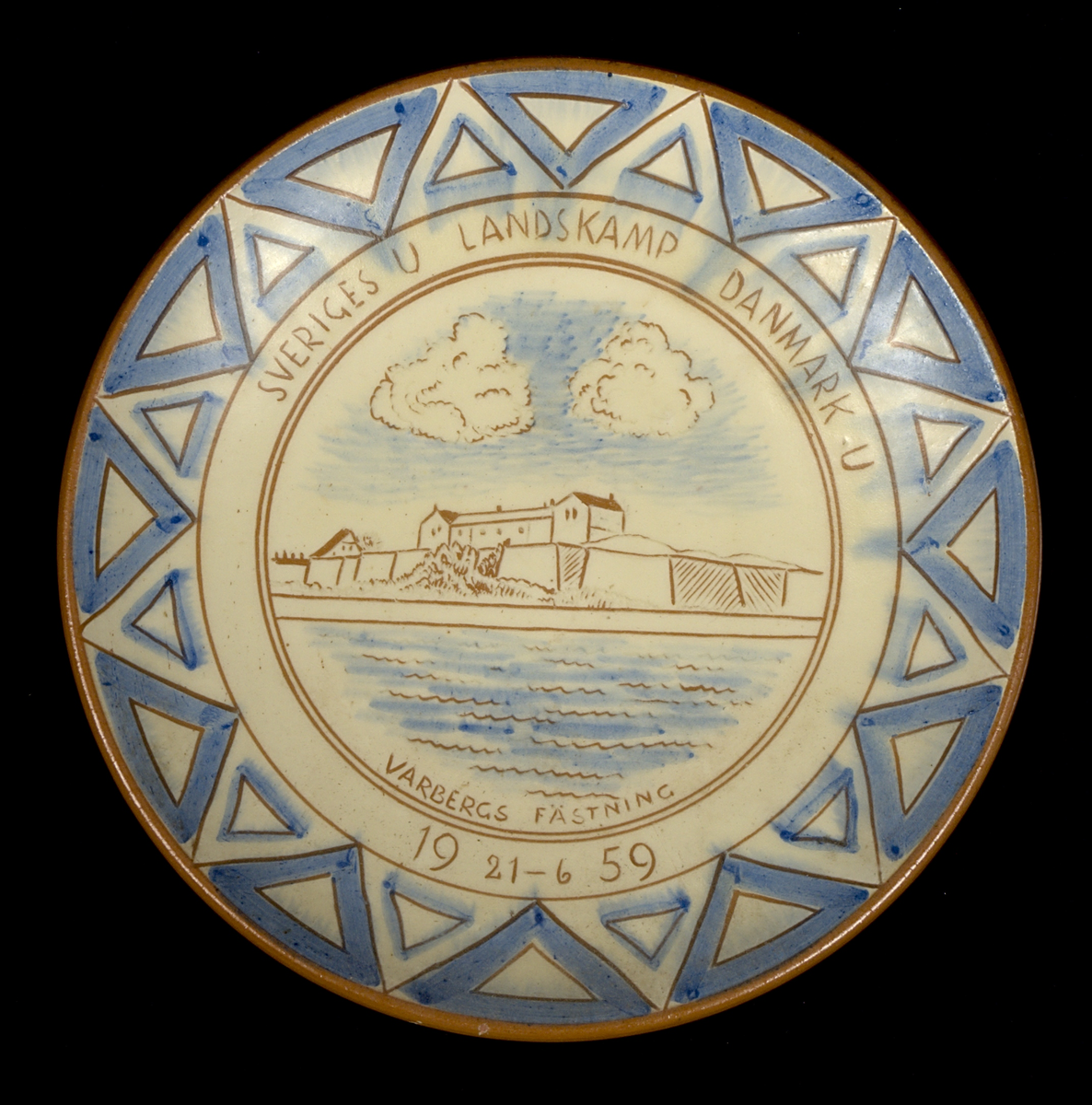 Runt fat av lergods med motiv i blått och brunt mot vit botten. Fatet är dekorerat med en geometriska bård längs kanten och med en vy av Varbergs fästning i spegeln. Text: "SVERIGES U LANDSKAMP DAMNARK U. VARBERGS FÄSTNING 19 21-6 59". På fatets undersida finns ristad signatur (se historik).