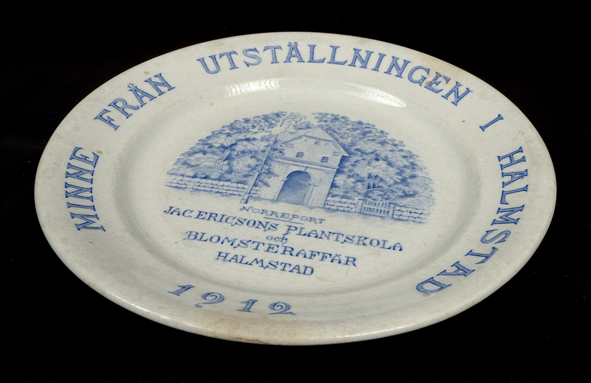 Prydnadsfat av flintgods med tryckt motiv i blått. Längs brämet löper texten: "MINNE FRÅN UTSTÄLLNINGEN I HALMSTAD 1912". I fatets spegel avbildas Norreport i Halmstad, samt texten: "JAC.ERICSONS PLANTSKOLA och BLOMSTERAFFÄR HALMSTAD". Undersidan är stämplad "Rörstrand".

Glasyr krackelerad.