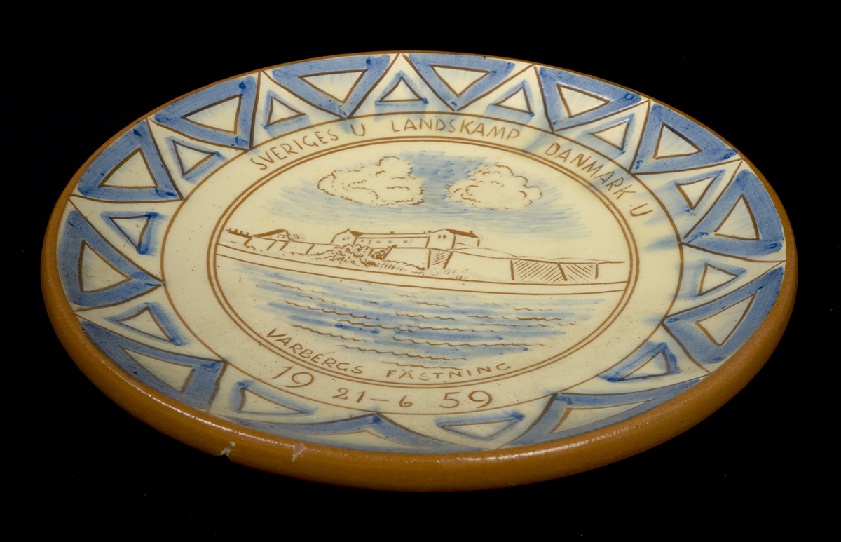 Runt fat av lergods med motiv i blått och brunt mot vit botten. Fatet är dekorerat med en geometriska bård längs kanten och med en vy av Varbergs fästning i spegeln. Text: "SVERIGES U LANDSKAMP DAMNARK U. VARBERGS FÄSTNING 19 21-6 59". På fatets undersida finns ristad signatur (se historik).