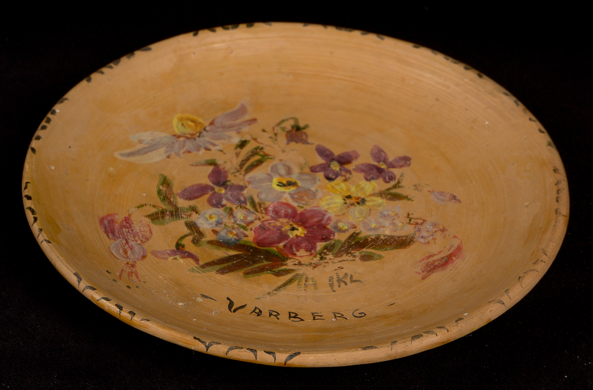 Prydnadsfat av lergods med handmålad blomsterdekor och texten "Varberg" i spegeln. Måleriet signerat: "IKL". Fatet är runt och svagt skålformat med låg kant. Tydliga drejspår samt fingeravtryck. Fatet bär en genomskinlig, mjölktonad glasyr, som ligger ovanpå dekoren. På undersidan av fatet finns en upphöjd fotring försedd med två hål för väggupphängning.

Signatur och årtal på undersidan: ”B(?)d.h. KIL. 75”. Även siffran ”3” skriven i blyerts.