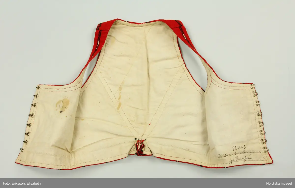 Huvudliggaren:
"Kvinnodräkt fr. Delsbo sn, Norra Helsinglands fgd, Helsingland.
a. "Ryntjeskjortel" af svart kläde med röd kant och bandväfd "kval" (linning). Kjolen "lurad"(goffrerad), skall förvaras lurad att ej vecken gå ut. 3 par fruntimmershänder hjälpas åt att "lura" den efter hvarje begagnande.
b. livstycke, rött med gröna ränder.
c. "Ryntjessärk" (rynksärk) af hvitt bomullstyg med "krus" (handbroderi) kring hals och händer.Begagnas sommartiden
d. "Bumsförkläde" af ylle, blårandigt med bandväf,"kval" i rödt och grönt.
e. "Förklesband" rödt och grönt ylle med "doppar" (tofsar) i ändarna. Lindas 1 à 2 hvarf kring lifvet och bindes framtill med stora nedhängande "locor" och ändar. Längd 262 cm, bredd 3,4 cm.
f. Kjolsäck med mässingslås och hake samt tennbroderi. Häktas under förklesbandet och hänger delvis under "försklet". Begagnas vanligen till förvaring af kyrkkryddor m.fl. kyrk-ellerkalassaker.
g. Halfhandskar af svart kläde med silkesbroderi. Begagnas sommartiden.
h. "Påshufva" hvardagsmössa af brokigt kattun. Begagnas med i nacken utslaget hår med några hårtestar hängande fritt framför öronen.
Till dräkten användes röda strumpor samt låga skor med höga klackar. Äfven kan användas silfersmycken kring halsen eller å  bröstet. En á två brokiga silkesdukar brukades omkring halsen nedstoppade under lifstycket, så att kruset kring halsen  på "ryntjessärken" sammanfästadt med ett löfspänne samt en smalare kant af hvarje sidenduk, blevo synliga. Vid handlederna sammanfästes ryntjessärken med röda sidenband eller länkknappar.

a. KJOL 
längd: 87 cm
midjeband bredd: 7 cm
av svart halvylle med svart linvarp och inslag av svart 1-tr. ullgarn, de 2 yttersta stadtrådarna är av oblekt lingarn. 4 goffrerade våder samt en slät framvåd som skarvats under förklädet med oblekt bomullslärft. 5 cm bred list av rött kläde i nederkanten runtom. Brett midjeband mönstervävt i rött och grönt ullgarn och vitt bomullsgarn. Knäppt vid framvådens vänstra sida med 2 hakar och hyskor av mässing.
b. LIVSTYCKE
längd: 39 cm
av halvylle i varptäckande kypert, varp av vitt lingarn, inslag av svart och grönt ullgarn med flaminslag av vitt, 2 fram-och sidstycken, 2 bakstycken avsmalnande till skörtveck med tre par tränsade förstärkningar av svart garn. I midjehöjd ett avsytt veck runtom, knäppt kant i kant med 8 par grova hakar och hyskor av mässing. Foder av ofärgad bomullslärft.
BE

c. ÖVERDEL
Längd: 39 cm. Längd: 58 cm - ärmlängd. Vidd: 56 cm - ärmvidd. Vidd: 114 cm. Höjd: 1,7 cm - kraghöjd. Material: Textil, bomull. Textil, linne - ärmlinning, krage. Bomullsgarn - broderi. Telnik: Tuskaft. Trådbundet broderi. Broderi, rätlinjig plattsöm. Handsömnad.
Överdel av tuskaftat vitt bomullstyg. Dubbelvikt bålstycke, isydda rektangulära halsspjäll. Täta breda stripade  rynkor mot halskrage i bomull, 1,7 cm hög. Kragen har broderier i rätlinjig plattsöm, vitt bomullsgarn över hela ytan, nuggor i kanten, langetterade bågar  i framkant. 16 cm långt sprund fram, tränsad förstärkning nedtill, 3 cm.  Vidsydd ärm med  infällda ärmspjäll, lagda veck över axeln fram och bak. Täta breda stripade rynkor mot 2,5 cm bred ärmlinning. Ärmlinningen har yttäckande vita broderier i rätlinjig plattsöm, rombiska former. Vita knytband i bomull i ärmlinningar och krage. Överdelen är handsydd.
/Inga-Lill Eliasson 2007-11-26


d. FÖRKLÄDE
längd=vådbredd: 82 cm
bredd: 97,5 cm
av halvylle, varp av svart lingarn, inslag i varptäckande kypert av svart 1-tr. ullgarn med smala ränder istarkt blått och en knappt synlig svart tråd med vita flamgarnsfläckar av bomull. Upptill stripade rynkor där de blå bårderna utnyttjats dekorativt i rynkningen. Mönstervävt midjeband i grönt på rött ullgarn , bandet knäpps med 2 hakar och hyskor i vänster sida.
I nederkantens båda hörn infodringar på insidan av svart bomullslärft, för att tyget inte skall rulla sig.



Berit Eldvik 2004