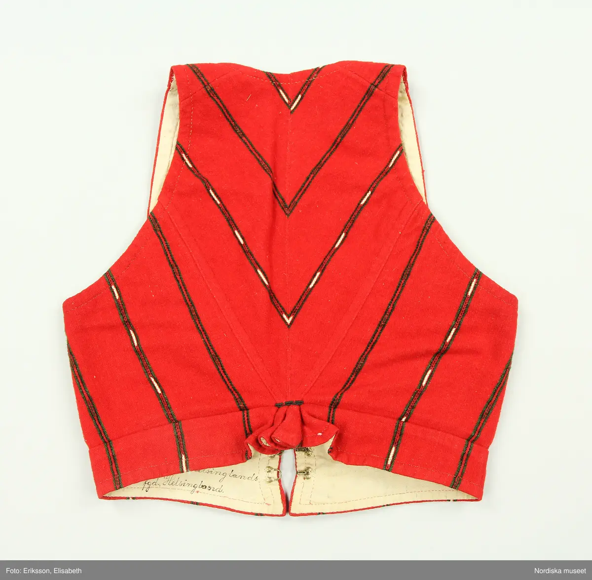 Huvudliggaren:
"Kvinnodräkt fr. Delsbo sn, Norra Helsinglands fgd, Helsingland.
a. "Ryntjeskjortel" af svart kläde med röd kant och bandväfd "kval" (linning). Kjolen "lurad"(goffrerad), skall förvaras lurad att ej vecken gå ut. 3 par fruntimmershänder hjälpas åt att "lura" den efter hvarje begagnande.
b. livstycke, rött med gröna ränder.
c. "Ryntjessärk" (rynksärk) af hvitt bomullstyg med "krus" (handbroderi) kring hals och händer.Begagnas sommartiden
d. "Bumsförkläde" af ylle, blårandigt med bandväf,"kval" i rödt och grönt.
e. "Förklesband" rödt och grönt ylle med "doppar" (tofsar) i ändarna. Lindas 1 à 2 hvarf kring lifvet och bindes framtill med stora nedhängande "locor" och ändar. Längd 262 cm, bredd 3,4 cm.
f. Kjolsäck med mässingslås och hake samt tennbroderi. Häktas under förklesbandet och hänger delvis under "försklet". Begagnas vanligen till förvaring af kyrkkryddor m.fl. kyrk-ellerkalassaker.
g. Halfhandskar af svart kläde med silkesbroderi. Begagnas sommartiden.
h. "Påshufva" hvardagsmössa af brokigt kattun. Begagnas med i nacken utslaget hår med några hårtestar hängande fritt framför öronen.
Till dräkten användes röda strumpor samt låga skor med höga klackar. Äfven kan användas silfersmycken kring halsen eller å  bröstet. En á två brokiga silkesdukar brukades omkring halsen nedstoppade under lifstycket, så att kruset kring halsen  på "ryntjessärken" sammanfästadt med ett löfspänne samt en smalare kant af hvarje sidenduk, blevo synliga. Vid handlederna sammanfästes ryntjessärken med röda sidenband eller länkknappar.

a. KJOL 
längd: 87 cm
midjeband bredd: 7 cm
av svart halvylle med svart linvarp och inslag av svart 1-tr. ullgarn, de 2 yttersta stadtrådarna är av oblekt lingarn. 4 goffrerade våder samt en slät framvåd som skarvats under förklädet med oblekt bomullslärft. 5 cm bred list av rött kläde i nederkanten runtom. Brett midjeband mönstervävt i rött och grönt ullgarn och vitt bomullsgarn. Knäppt vid framvådens vänstra sida med 2 hakar och hyskor av mässing.
b. LIVSTYCKE
längd: 39 cm
av halvylle i varptäckande kypert, varp av vitt lingarn, inslag av svart och grönt ullgarn med flaminslag av vitt, 2 fram-och sidstycken, 2 bakstycken avsmalnande till skörtveck med tre par tränsade förstärkningar av svart garn. I midjehöjd ett avsytt veck runtom, knäppt kant i kant med 8 par grova hakar och hyskor av mässing. Foder av ofärgad bomullslärft.
BE

c. ÖVERDEL
Längd: 39 cm. Längd: 58 cm - ärmlängd. Vidd: 56 cm - ärmvidd. Vidd: 114 cm. Höjd: 1,7 cm - kraghöjd. Material: Textil, bomull. Textil, linne - ärmlinning, krage. Bomullsgarn - broderi. Telnik: Tuskaft. Trådbundet broderi. Broderi, rätlinjig plattsöm. Handsömnad.
Överdel av tuskaftat vitt bomullstyg. Dubbelvikt bålstycke, isydda rektangulära halsspjäll. Täta breda stripade  rynkor mot halskrage i bomull, 1,7 cm hög. Kragen har broderier i rätlinjig plattsöm, vitt bomullsgarn över hela ytan, nuggor i kanten, langetterade bågar  i framkant. 16 cm långt sprund fram, tränsad förstärkning nedtill, 3 cm.  Vidsydd ärm med  infällda ärmspjäll, lagda veck över axeln fram och bak. Täta breda stripade rynkor mot 2,5 cm bred ärmlinning. Ärmlinningen har yttäckande vita broderier i rätlinjig plattsöm, rombiska former. Vita knytband i bomull i ärmlinningar och krage. Överdelen är handsydd.
/Inga-Lill Eliasson 2007-11-26


d. FÖRKLÄDE
längd=vådbredd: 82 cm
bredd: 97,5 cm
av halvylle, varp av svart lingarn, inslag i varptäckande kypert av svart 1-tr. ullgarn med smala ränder istarkt blått och en knappt synlig svart tråd med vita flamgarnsfläckar av bomull. Upptill stripade rynkor där de blå bårderna utnyttjats dekorativt i rynkningen. Mönstervävt midjeband i grönt på rött ullgarn , bandet knäpps med 2 hakar och hyskor i vänster sida.
I nederkantens båda hörn infodringar på insidan av svart bomullslärft, för att tyget inte skall rulla sig.



Berit Eldvik 2004