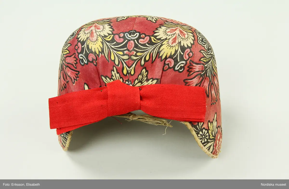 Huvudliggaren:
"Kvinnodräkt fr. Delsbo sn, Norra Helsinglands fgd, Helsingland.
a. "Ryntjeskjortel" af svart kläde med röd kant och bandväfd "kval" (linning). Kjolen "lurad"(goffrerad), skall förvaras lurad att ej vecken gå ut. 3 par fruntimmershänder hjälpas åt att "lura" den efter hvarje begagnande.
b. livstycke, rött med gröna ränder.
c. "Ryntjessärk" (rynksärk) af hvitt bomullstyg med "krus" (handbroderi) kring hals och händer.Begagnas sommartiden
d. "Bumsförkläde" af ylle, blårandigt med bandväf,"kval" i rödt och grönt.
e. "Förklesband" rödt och grönt ylle med "doppar" (tofsar) i ändarna. Lindas 1 à 2 hvarf kring lifvet och bindes framtill med stora nedhängande "locor" och ändar. Längd 262 cm, bredd 3,4 cm.
f. Kjolsäck med mässingslås och hake samt tennbroderi. Häktas under förklesbandet och hänger delvis under "försklet". Begagnas vanligen till förvaring af kyrkkryddor m.fl. kyrk-ellerkalassaker.
g. Halfhandskar af svart kläde med silkesbroderi. Begagnas sommartiden.
h. "Påshufva" hvardagsmössa af brokigt kattun. Begagnas med i nacken utslaget hår med några hårtestar hängande fritt framför öronen.
Till dräkten användes röda strumpor samt låga skor med höga klackar. Äfven kan användas silfersmycken kring halsen eller å  bröstet. En á två brokiga silkesdukar brukades omkring halsen nedstoppade under lifstycket, så att kruset kring halsen  på "ryntjessärken" sammanfästadt med ett löfspänne samt en smalare kant af hvarje sidenduk, blevo synliga. Vid handlederna sammanfästes ryntjessärken med röda sidenband eller länkknappar.