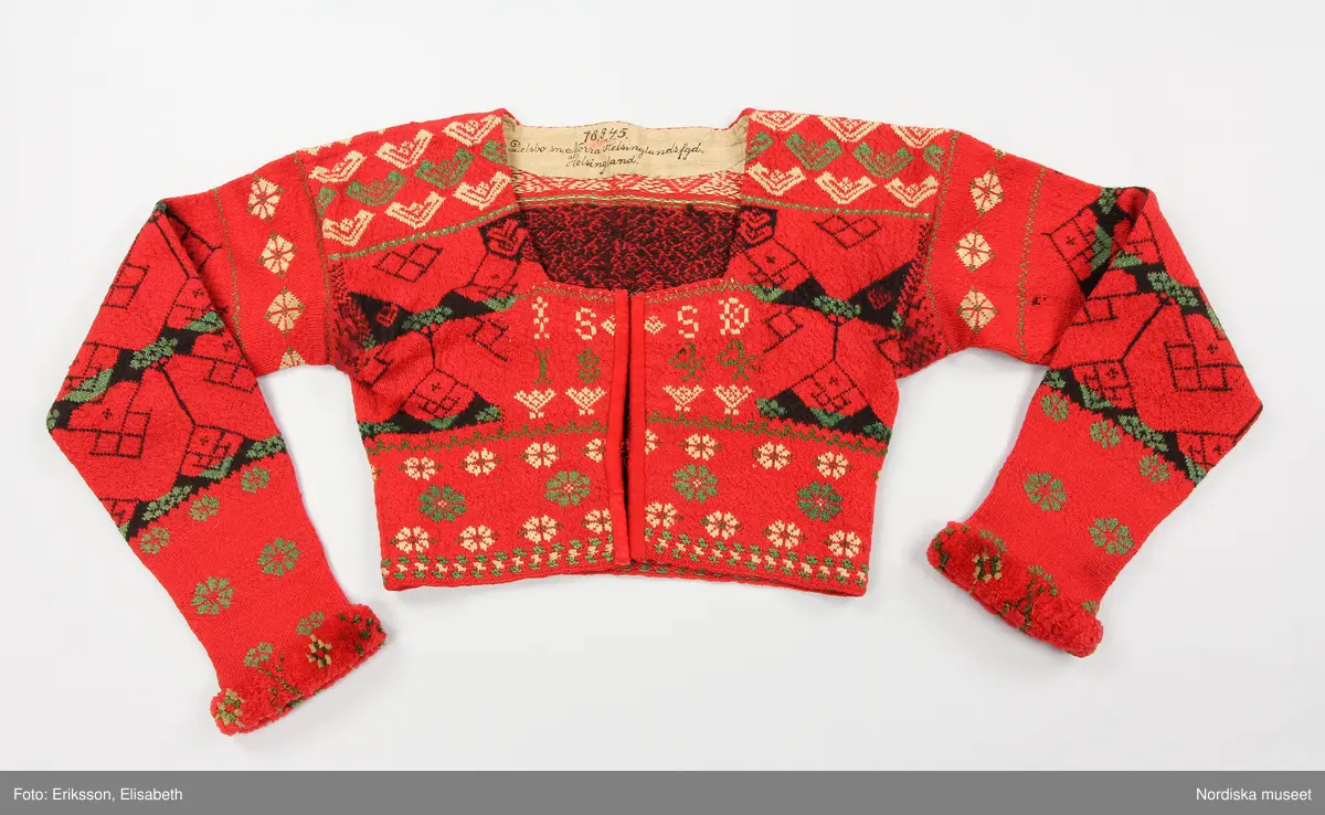 Kvinnotröja, midjekort, slätstickad i hårt tvinnat tunt ullgarn, helt mönstrad i rött som dominant färg, samt svart, grönt och vitt. Det vita garnet är bomullsgarn, vilket föredrogs för att det gav en renare vit färg än ullgarn. Mönstret är specifikt för socknarna Delsbo och Bjuråker. Stickningen utförd så att trådarna binder hela vägen på baksidan. Bålen rundstickad och sedan uppklippt mitt fram, där öppningen kantats med rött kläde. Fyrkantig halsringning infodrad med oblekt linnelärft. Knäppt med 5 par hakar och hyskor av mässing. Ärmarna stickade för sig och vidsydda mot bålen. Ärmarna avsmalnar mot handleden där de kantats med ett flossabräm av  ullgarn i rött med mönster i grönt och vitt. Brämet vävt med botten av lingarn och inknutna nockor som sedan klippts kort och jämnt.
På bröstet instickad märkning I S S D  1 8 4 4 = jungfru Sigrid Svensdotter.
Dessa tröjor blev moderna kring 1830-40 tal i Delsbo och användes som söndagseftermiddagsdräkt framförallt av de unga som då samlades till dans eller andra nöjen. Männens tröjor var av samma slag men var inte öppna fram.
/Berit Eldvik 2007-07-03