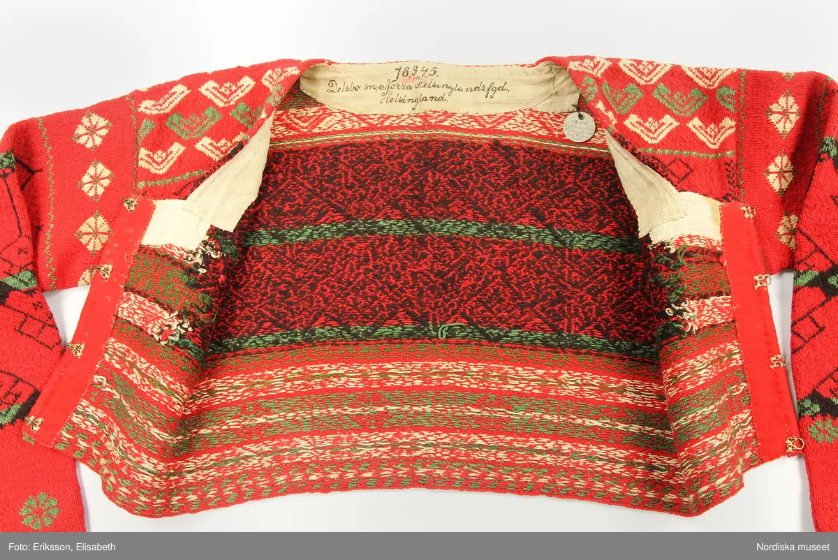 Kvinnotröja, midjekort, slätstickad i hårt tvinnat tunt ullgarn, helt mönstrad i rött som dominant färg, samt svart, grönt och vitt. Det vita garnet är bomullsgarn, vilket föredrogs för att det gav en renare vit färg än ullgarn. Mönstret är specifikt för socknarna Delsbo och Bjuråker. Stickningen utförd så att trådarna binder hela vägen på baksidan. Bålen rundstickad och sedan uppklippt mitt fram, där öppningen kantats med rött kläde. Fyrkantig halsringning infodrad med oblekt linnelärft. Knäppt med 5 par hakar och hyskor av mässing. Ärmarna stickade för sig och vidsydda mot bålen. Ärmarna avsmalnar mot handleden där de kantats med ett flossabräm av  ullgarn i rött med mönster i grönt och vitt. Brämet vävt med botten av lingarn och inknutna nockor som sedan klippts kort och jämnt.
På bröstet instickad märkning I S S D  1 8 4 4 = jungfru Sigrid Svensdotter.
Dessa tröjor blev moderna kring 1830-40 tal i Delsbo och användes som söndagseftermiddagsdräkt framförallt av de unga som då samlades till dans eller andra nöjen. Männens tröjor var av samma slag men var inte öppna fram.
/Berit Eldvik 2007-07-03