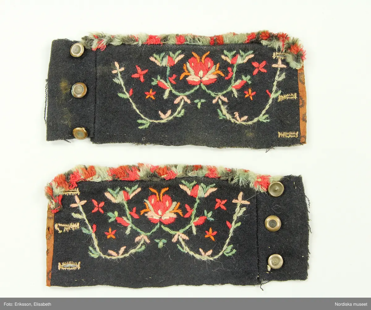 Av svart vadmal med randigt halvyllefoder. Broderi i plattsöm och stjälksöm med 1-tr. ullgarn i rött, rosa, orange, grönt och ljusblått, kantade på kortsidorna med ett tryckt brunt bomullstyg. Knäppt med 3 mässingsknappar och tränsade knapphål, knapparna fästade med skinnremsa på baksidan. Flossabräm av flera rader påsydd kavelfrans.

Berit Eldvik 2004