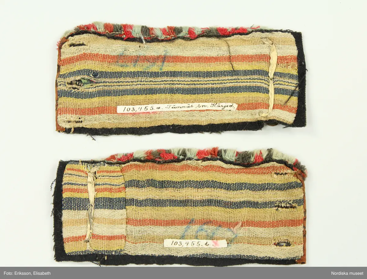 Av svart vadmal med randigt halvyllefoder. Broderi i plattsöm och stjälksöm med 1-tr. ullgarn i rött, rosa, orange, grönt och ljusblått, kantade på kortsidorna med ett tryckt brunt bomullstyg. Knäppt med 3 mässingsknappar och tränsade knapphål, knapparna fästade med skinnremsa på baksidan. Flossabräm av flera rader påsydd kavelfrans.

Berit Eldvik 2004