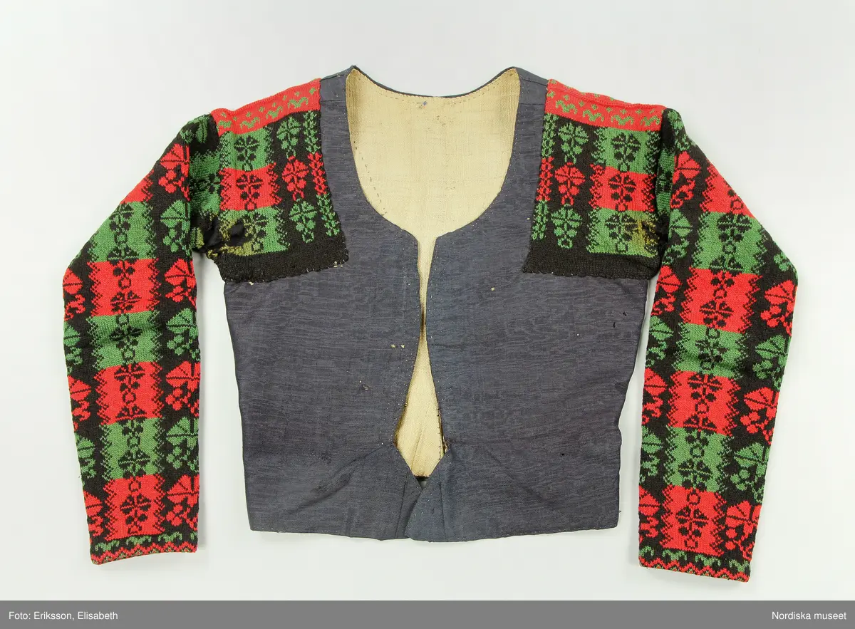 Ursprungligen ett livstycke av mörkblått kamgarnsylle i tuskaft med vattrad yta, s.k. kamlott. Livstycket i 1700-talssnitt med långt skört.
2 fram-och sidstycken, 2 bakstycken med 3 skörtveck i ryggen, 
Knäppning med 6 par hakar och hyskor, de av järntråd är äldst och ursprungliga, de i mässing yngre. Foder av grov oblekt linnelärft. Vid omsyning till tröja har skörtvecken sytts in i sömmarna ev. har hela livstycket även kortats något. Ärmarna mönsterstickade av rött tvinnat ullgarn och vitt bomullsgarn i stora rutor med nejlikmönster instickade i motsatsfärg. Ett rakt axelstycke stickat i samma mönstring och fastsytt vid ärmen och därefter påsytt på livstycket, så att det ursprungliga ärmhålet finns kvar under det stickade.
Anm. Ärmarna har fållats upp ca 2 cm

Kamlott var modetyg under senare delen av 1700-talet, det var ett dyrbart köpetyg och användes av allmogen i de finaste bröllopsplaggen som livstycken och kjol. Se t.ex svart kjol och tröja från Arbrå inv.nr 1824 och 1825. Ofta ser man sådana plagg omsydda till modernare snitt under 1800-talet. Ett så dyrbart och fint tyg återbrukades gärna. Jfr. liknande tröjor inv.nr 111497 och 201427 samt lösa ärmar 113011.
Troligen är samtliga dessa lösärmar stickade av samma person.
Beskrivning finns i boken Svenska Tröjor av Britt-Marie Christoffersson, ICA förlag 1988.
/Berit Eldvik 2006-06-16