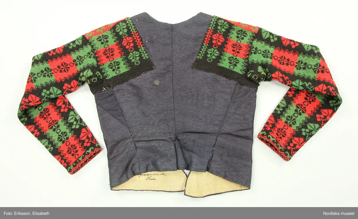 Ursprungligen ett livstycke av mörkblått kamgarnsylle i tuskaft med vattrad yta, s.k. kamlott. Livstycket i 1700-talssnitt med långt skört.
2 fram-och sidstycken, 2 bakstycken med 3 skörtveck i ryggen, 
Knäppning med 6 par hakar och hyskor, de av järntråd är äldst och ursprungliga, de i mässing yngre. Foder av grov oblekt linnelärft. Vid omsyning till tröja har skörtvecken sytts in i sömmarna ev. har hela livstycket även kortats något. Ärmarna mönsterstickade av rött tvinnat ullgarn och vitt bomullsgarn i stora rutor med nejlikmönster instickade i motsatsfärg. Ett rakt axelstycke stickat i samma mönstring och fastsytt vid ärmen och därefter påsytt på livstycket, så att det ursprungliga ärmhålet finns kvar under det stickade.
Anm. Ärmarna har fållats upp ca 2 cm

Kamlott var modetyg under senare delen av 1700-talet, det var ett dyrbart köpetyg och användes av allmogen i de finaste bröllopsplaggen som livstycken och kjol. Se t.ex svart kjol och tröja från Arbrå inv.nr 1824 och 1825. Ofta ser man sådana plagg omsydda till modernare snitt under 1800-talet. Ett så dyrbart och fint tyg återbrukades gärna. Jfr. liknande tröjor inv.nr 111497 och 201427 samt lösa ärmar 113011.
Troligen är samtliga dessa lösärmar stickade av samma person.
Beskrivning finns i boken Svenska Tröjor av Britt-Marie Christoffersson, ICA förlag 1988.
/Berit Eldvik 2006-06-16