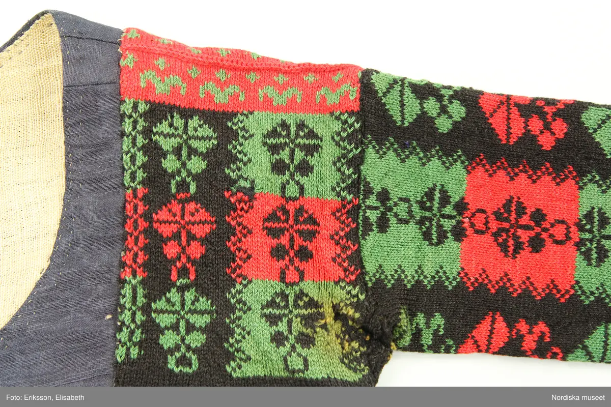 Ursprungligen ett livstycke av mörkblått kamgarnsylle i tuskaft med vattrad yta, s.k. kamlott. Livstycket i 1700-talssnitt med långt skört.
2 fram-och sidstycken, 2 bakstycken med 3 skörtveck i ryggen, 
Knäppning med 6 par hakar och hyskor, de av järntråd är äldst och ursprungliga, de i mässing yngre. Foder av grov oblekt linnelärft. Vid omsyning till tröja har skörtvecken sytts in i sömmarna ev. har hela livstycket även kortats något. Ärmarna mönsterstickade av rött tvinnat ullgarn och vitt bomullsgarn i stora rutor med nejlikmönster instickade i motsatsfärg. Ett rakt axelstycke stickat i samma mönstring och fastsytt vid ärmen och därefter påsytt på livstycket, så att det ursprungliga ärmhålet finns kvar under det stickade.
Anm. Ärmarna har fållats upp ca 2 cm

Kamlott var modetyg under senare delen av 1700-talet, det var ett dyrbart köpetyg och användes av allmogen i de finaste bröllopsplaggen som livstycken och kjol. Se t.ex svart kjol och tröja från Arbrå inv.nr 1824 och 1825. Ofta ser man sådana plagg omsydda till modernare snitt under 1800-talet. Ett så dyrbart och fint tyg återbrukades gärna. Jfr. liknande tröjor inv.nr 111497 och 201427 samt lösa ärmar 113011.
Troligen är samtliga dessa lösärmar stickade av samma person.
Beskrivning finns i boken Svenska Tröjor av Britt-Marie Christoffersson, ICA förlag 1988.
/Berit Eldvik 2006-06-16