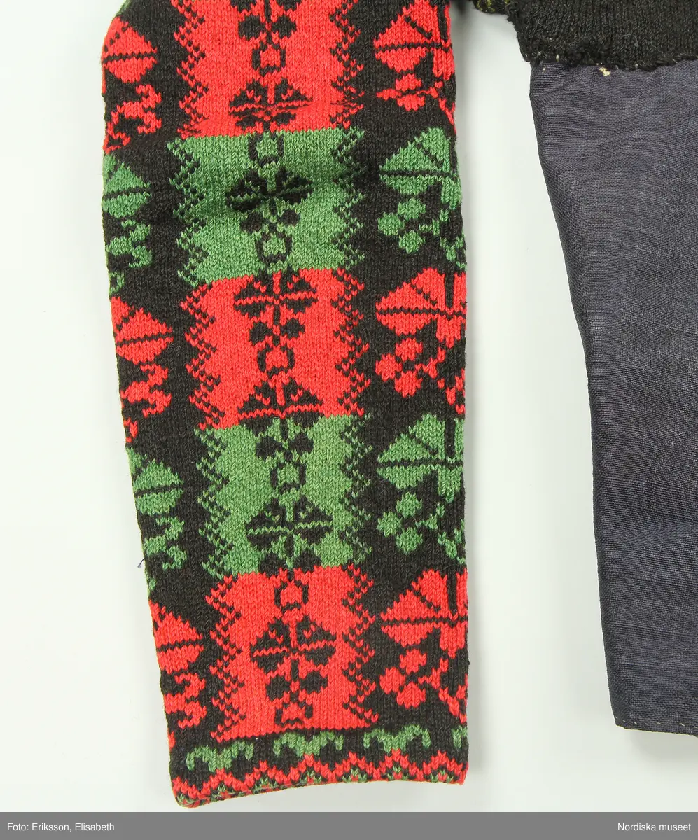 Ursprungligen ett livstycke av mörkblått kamgarnsylle i tuskaft med vattrad yta, s.k. kamlott. Livstycket i 1700-talssnitt med långt skört.
2 fram-och sidstycken, 2 bakstycken med 3 skörtveck i ryggen, 
Knäppning med 6 par hakar och hyskor, de av järntråd är äldst och ursprungliga, de i mässing yngre. Foder av grov oblekt linnelärft. Vid omsyning till tröja har skörtvecken sytts in i sömmarna ev. har hela livstycket även kortats något. Ärmarna mönsterstickade av rött tvinnat ullgarn och vitt bomullsgarn i stora rutor med nejlikmönster instickade i motsatsfärg. Ett rakt axelstycke stickat i samma mönstring och fastsytt vid ärmen och därefter påsytt på livstycket, så att det ursprungliga ärmhålet finns kvar under det stickade.
Anm. Ärmarna har fållats upp ca 2 cm

Kamlott var modetyg under senare delen av 1700-talet, det var ett dyrbart köpetyg och användes av allmogen i de finaste bröllopsplaggen som livstycken och kjol. Se t.ex svart kjol och tröja från Arbrå inv.nr 1824 och 1825. Ofta ser man sådana plagg omsydda till modernare snitt under 1800-talet. Ett så dyrbart och fint tyg återbrukades gärna. Jfr. liknande tröjor inv.nr 111497 och 201427 samt lösa ärmar 113011.
Troligen är samtliga dessa lösärmar stickade av samma person.
Beskrivning finns i boken Svenska Tröjor av Britt-Marie Christoffersson, ICA förlag 1988.
/Berit Eldvik 2006-06-16