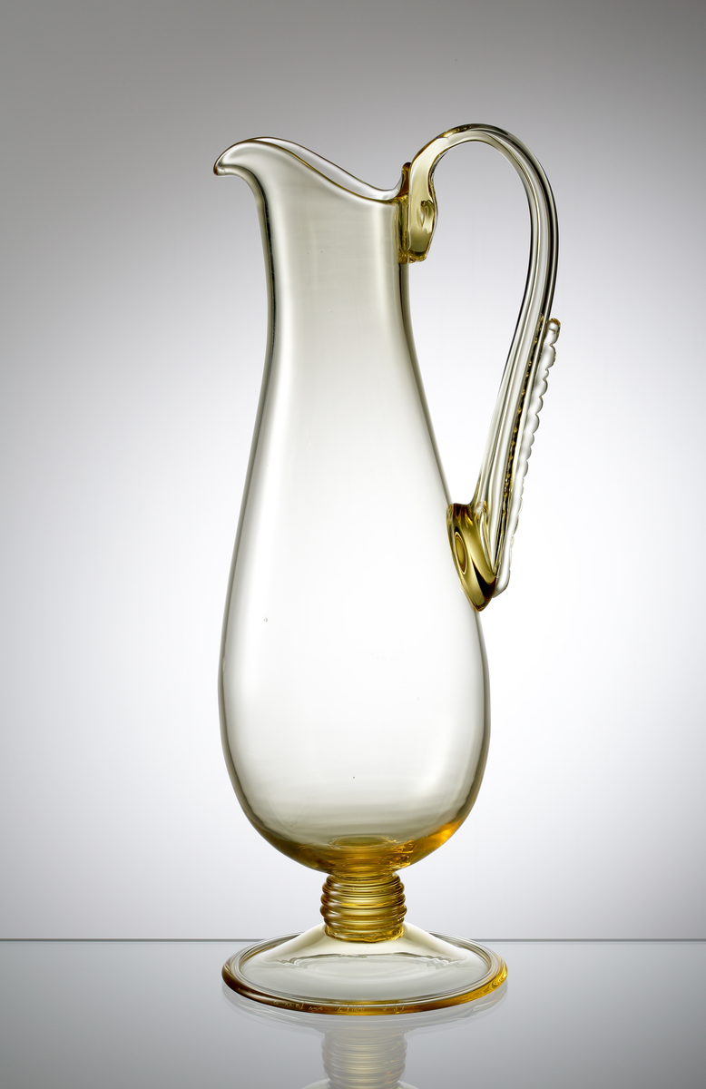 Formgivare: Nils Landberg. Kanna på fot. Expo. Utförd i bärnstenstonat glas. Droppformad kropp med utdragen snip. Grepe med luft samt pålagd nupen tråd. Lågt räfflat ben. Utvikt fot med omvikt kant.