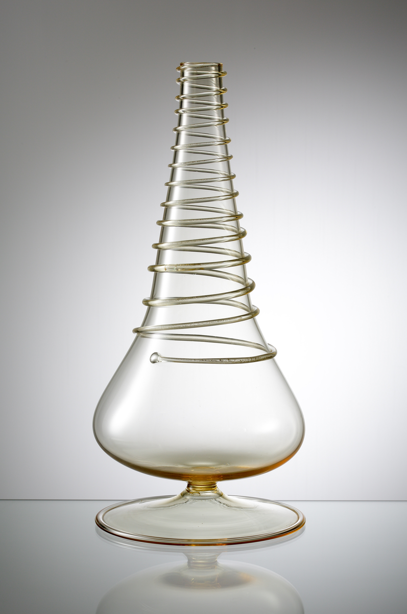 Formgivare: Nils Landberg. Vas på fot. Expo. Utförd i bärnstenstonat glas. Droppformad kropp med pålagd glastråd i spiral. Utsvängd fot med omvikt kant.