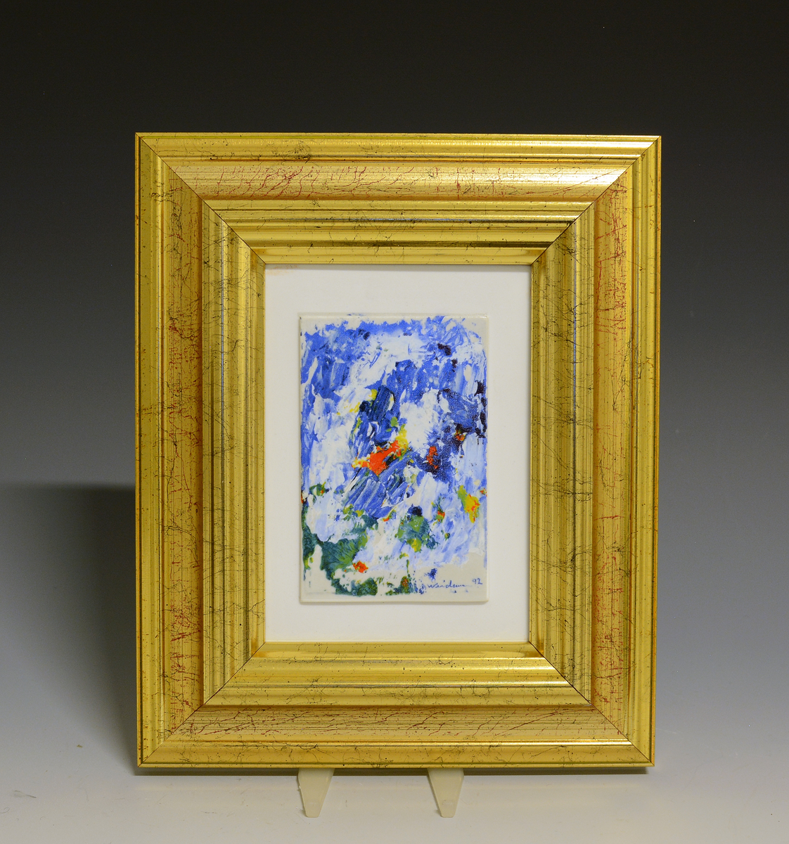 Porselensbilde med bred gullramme. Abstrakt motiv i blått, grønt, gult og rødt. 
1203/2500