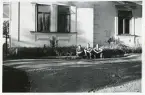 Tre personer som sitter på marken framför ett hus. Text baksida kort: Östanby 1947.