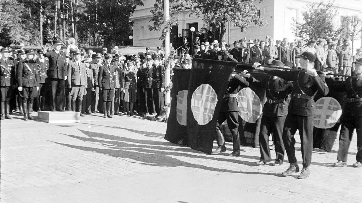 Et historisk svart-hvitt-fotografi fra et Nasjonal Samling (NS)-arrangement i Norge på 1940-tallet. Til venstre står en mann i mørk uniform, sannsynligvis Vidkun Quisling, på en lav trepall og gjør en hilsen. I forgrunnen marsjerer flere menn i mørke uniformer forbi mens de bærer store, svarte flagg med det hvite solkors-symbolet. En folkemengde av uniformerte tjenestemenn og sivile tilskuere står i bakgrunnen foran en stor, lys klassisistisk bygning. Scenen utspiller seg utendørs på et brosteinsbelagt område på dagtid.