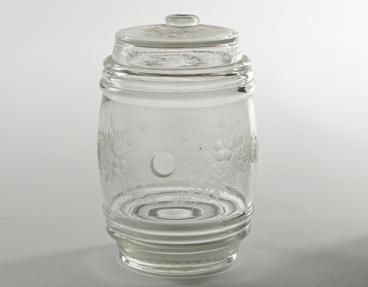 Tønneformet glassbeholder med lokk. Det er skåret ut hull i sideveggen. Dekorasjon av blomster og blader er slepet rundt hele korpus og kanten på lokket.