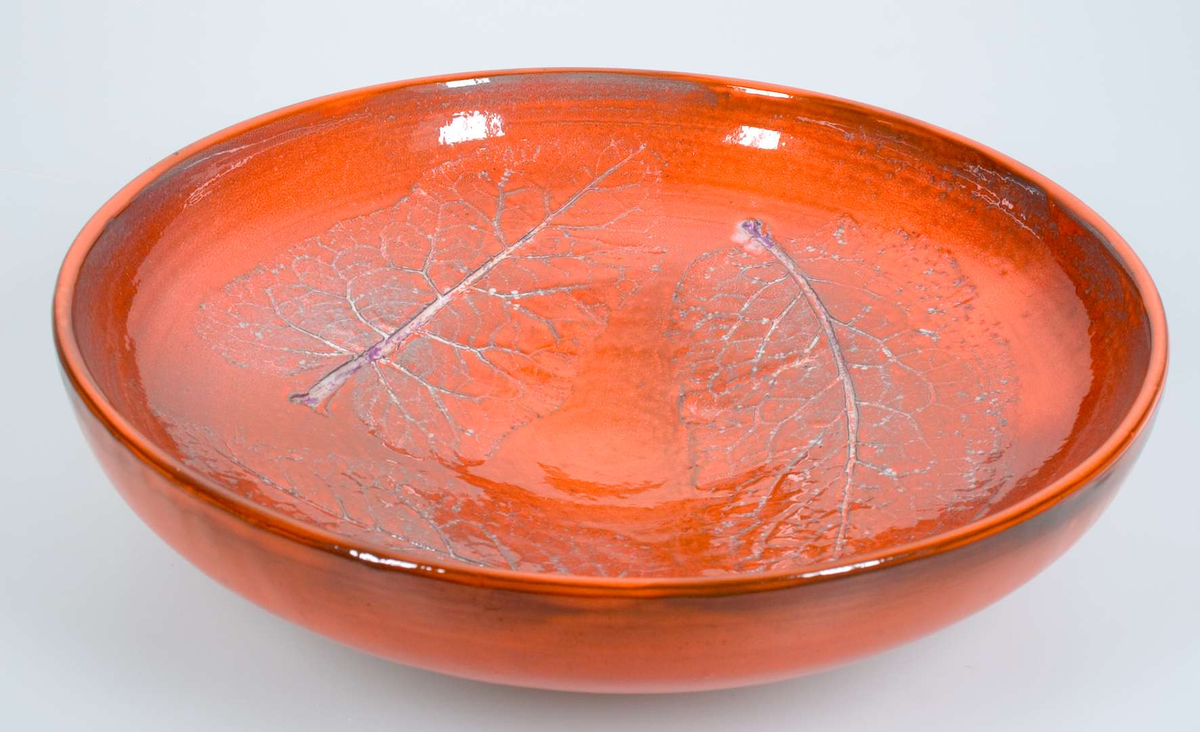 Av lergods mönster tryckt i massan med tussilagoblad. orange överglasyr med ljusgrå underglasyr. Signerad ABG 1970.