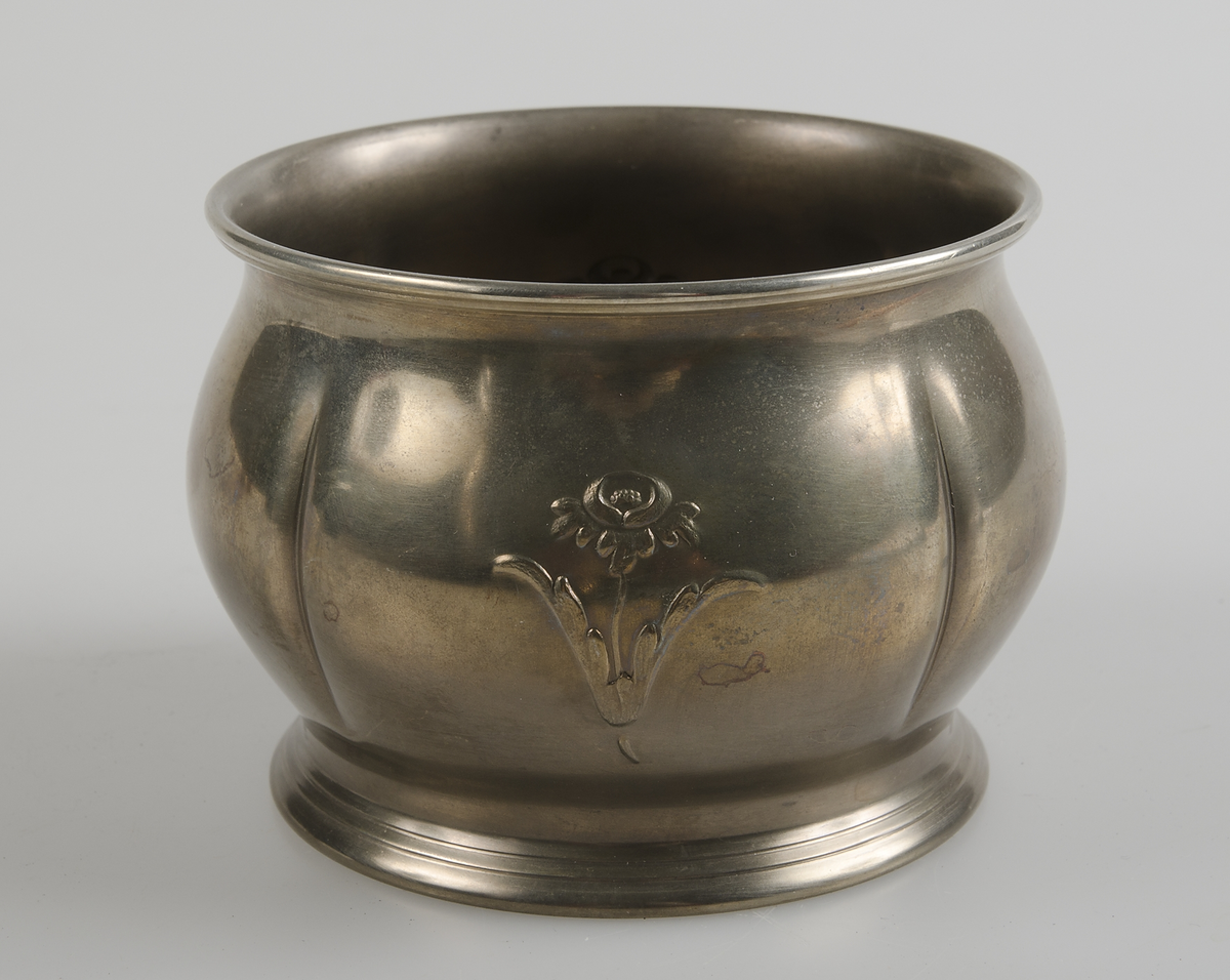 Del till sockerskål, rundel av gul metall med två blommor i relief. Botten och handtag saknas.
Sockerskål nodell nr 27 i katalog nr 20.